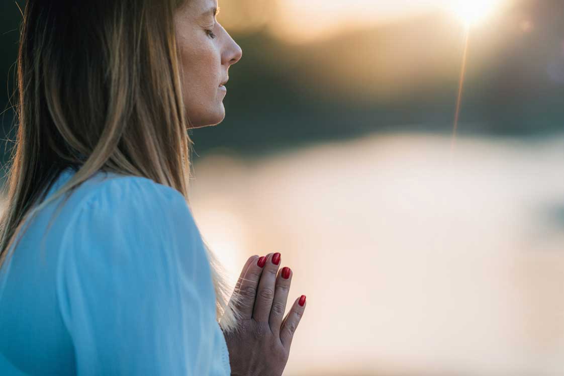 Woman with closed eyes and hands together in prayer at sunset.